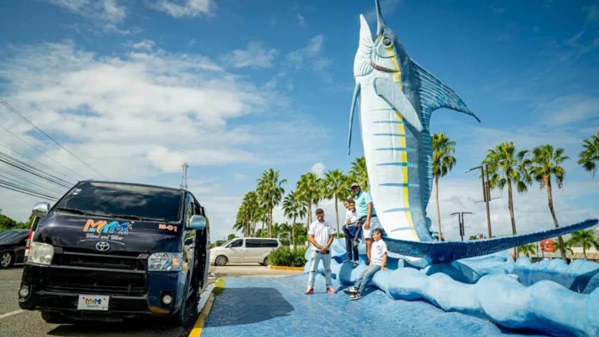 Una van rotulada de MyM Travel & Tours junto a la escultura del pez marlin azul, con palmeras al fondo